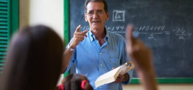 Menina levantando a mão fazendo perguntas ao professor na escola. Foto que ilustra post sobre políticas públicas municipais. Reprodução: diego_cervo/Envato.
