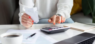 Foto ilustrando redução de custos operacionais, com mulher fazendo contas usando calculadora e computador