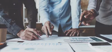 Pessoas em reunião, com calculadora e papéis espalhados na mesa. Foto que ilustra post sobre economia de escala em projetos públicos.