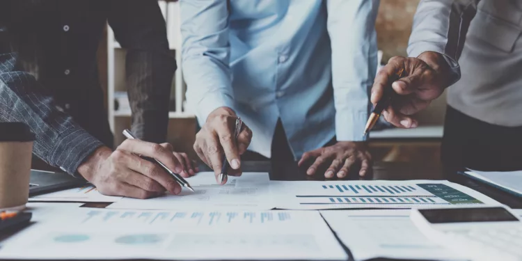 Pessoas em reunião, com calculadora e papéis espalhados na mesa. Foto que ilustra post sobre economia de escala em projetos públicos.