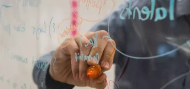 Pessoa usando quadro transparente para escrever ideias, em foto que ilustra post sobre Lei de Inovação Tecnológica. Reprodução: Kvalifik/Unsplash