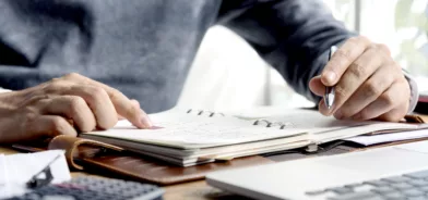 Pessoa trabalhando, usando computador e agenda, ilustrando post sobre Plano de Contratações Anual. Reprodução: Rawpixel/Envato.