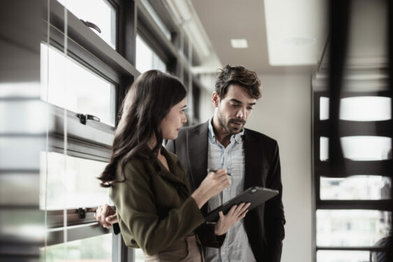 Duas pessoas em um ambiente de trabalho, discutindo algo enquanto observam um tablet, em frente a uma janela grande. Foto que ilustra post sobre governança corporativa na gestão pública. SpaceOak/Envato.