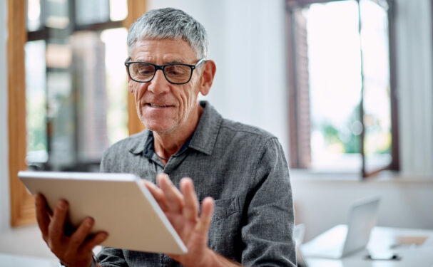 Homem idoso de cabelos grisalhos e óculos, segurando um tablet com uma expressão concentrada e leve sorriso. Foto que ilustra post sobre acessibilidade digital. Reprodução: YuriArcursPeopleimages/Envato.