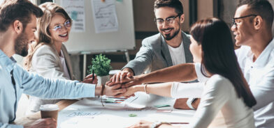 Grupo de pessoas em uma reunião de trabalho, todos sorrindo e com as mãos juntas no centro da mesa, em um gesto de união e colaboração. Foto ilustra post sobre gestão integrada. Reprodução: gstockstudio/Envato.