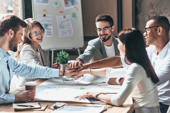 Grupo de pessoas em uma reunião de trabalho, todos sorrindo e com as mãos juntas no centro da mesa, em um gesto de união e colaboração. Foto ilustra post sobre gestão integrada. Reprodução: gstockstudio/Envato.