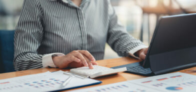 Pessoa trabalhando em um laptop, usando uma calculadora e analisando documentos que contêm gráficos e dados estatísticos. Foto que ilustra post sobre políticas públicas baseadas em evidências. Reprodução: AmnajKhetsamtip/Envato.