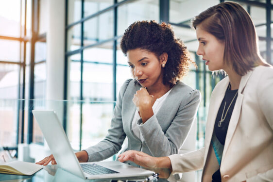 Duas mulheres em um ambiente de escritório moderno, olhando atentamente para a tela de um laptop. Foto que ilustra post sobre produtividade. Reprodução: YuriArcursPeopleimages/Envato.