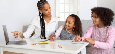 Uma mãe está ensinando duas meninas pequenas em casa, usando um laptop e materiais de escrita, enquanto aponta para a tela. Foto que ilustra post sobre alfabetização digital. Reprodução: Prostock-studio/Envato.