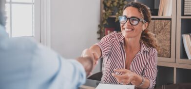 Uma mulher sorridente, usando óculos e camisa listrada, aperta a mão de outra pessoa em um ambiente de escritório. Foto que ilustra post sobre boas práticas de governança pública. Reprodução: PerfectWave003/Envato.