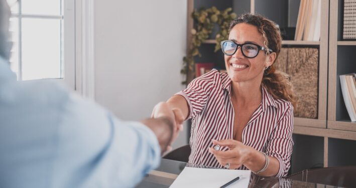 Uma mulher sorridente, usando óculos e camisa listrada, aperta a mão de outra pessoa em um ambiente de escritório. Foto que ilustra post sobre boas práticas de governança pública. Reprodução: PerfectWave003/Envato.