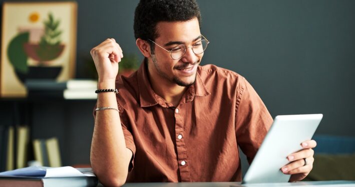 Jovem estudante sorridente durante estudo on-line olhando para a tela do tablet. Foto que ilustra post sobre educação digital. Reprodução: Pressmaster/Envato.