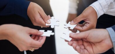 Na imagem, várias mãos seguram peças de um quebra-cabeça, simbolizando colaboração e integração de equipes para alcançar uma solução conjunta. Reprodução: YuriArcursPeopleimages/Envato.