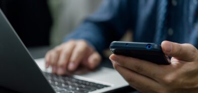 Uma pessoa está usando um smartphone enquanto digita em um laptop, em foto que ilustra post sobre TIC. Reprodução: donut3771/Envato.