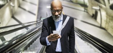 Empresário com smartphone lendo mensagens na escada rolante, em foto que ilustra post sobre atendimento ao cidadão. Reprodução: westend61/Envato.