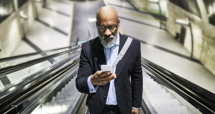 Empresário com smartphone lendo mensagens na escada rolante, em foto que ilustra post sobre atendimento ao cidadão. Reprodução: westend61/Envato.