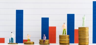 Figuras de pessoas em moedas na superfície branca com gráficos ao fundo. Foto que ilustra post sobre princípio da economicidade. Reprodução: LightFieldStudios/Envato.