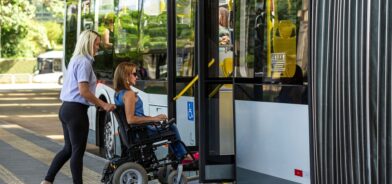 Mulher em cadeira de rodas embarca em ônibus acessível com auxílio de outra pessoa, em foto que ilustra post sobre serviços essenciais. Reprodução: wirestock/Envato.
