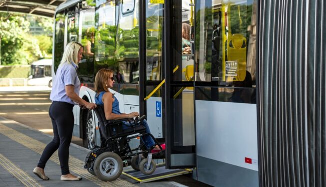 Mulher em cadeira de rodas embarca em ônibus acessível com auxílio de outra pessoa, em foto que ilustra post sobre serviços essenciais. Reprodução: wirestock/Envato.