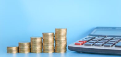 Pilha de moedas de dinheiro em fundo azul com calculadora. Foto que ilustra post sobre sustentabilidade fiscal. Reprodução: tendo23/Envato.