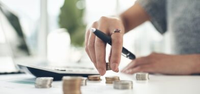 Pessoa contando moedas e fazendo conta em calculadora. Foto que ilustra post sobre dívida ativa municipal. Reprodução: YuriArcursPeopleimages/Envato.