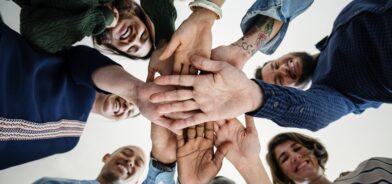 Grupo de pessoas em círculo, vistas de baixo para cima, sorrindo e com as mãos sobrepostas no centro, ilustrando post sobre Organização da Sociedade Civil (OCS). Reprodução: Rawpixel/Envato.