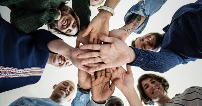Grupo de pessoas em círculo, vistas de baixo para cima, sorrindo e com as mãos sobrepostas no centro, ilustrando post sobre Organização da Sociedade Civil (OCS). Reprodução: Rawpixel/Envato.