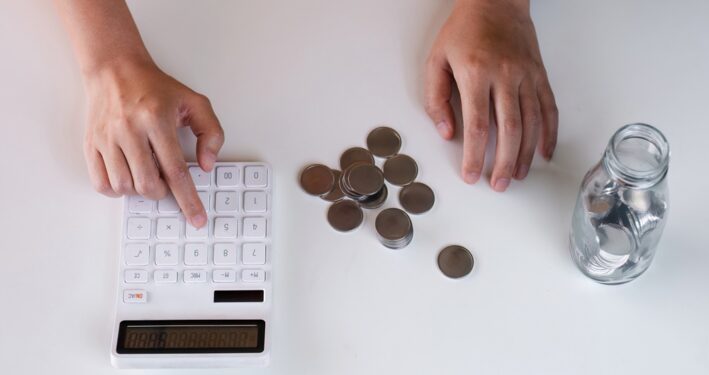 Pessoa contando moedas e fazendo contas em calculadora. Foto que ilustra post sobre Receita Corrente Líquida (RCL). Reprodução: wichayada69/Envato.