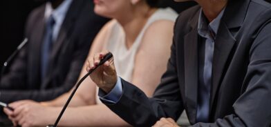 Parlamentar ajustando microfone discutindo legislação com colegas durante reunião. Foto que ilustra post sobre comissão parlamentar. Reprodução: seventyfourimages/Envato.