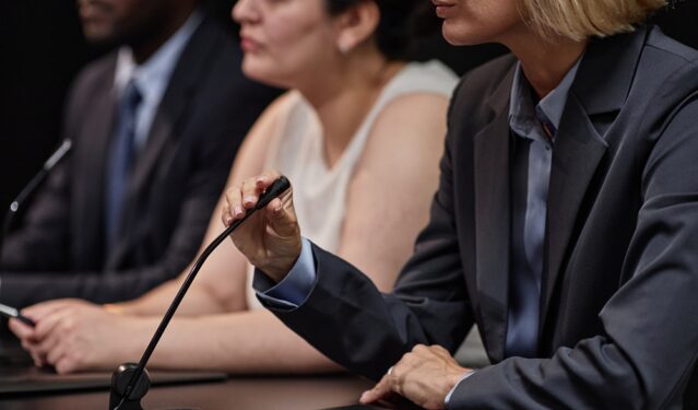 Parlamentar ajustando microfone discutindo legislação com colegas durante reunião. Foto que ilustra post sobre comissão parlamentar. Reprodução: seventyfourimages/Envato.