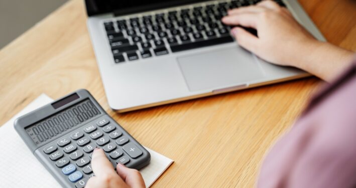 Pessoa usando calculadora e computador ao mesmo tempo. Foto que ilustra post sobre DAIR nos RPPS. Reprodução: OaklandImages/Envato.