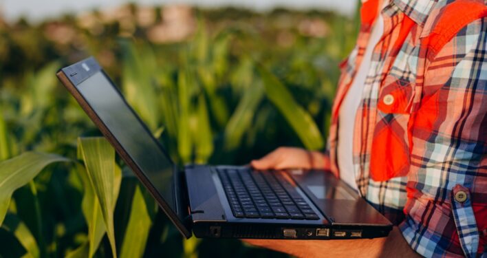 Agricultor segurando computador no campo. Foto que ilustra post sobre agricultura inteligente. Reprodução: SkloStudio/Envato.