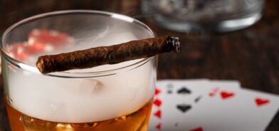 Charuto cubano, copo de uísque e cartas na mesa. Foto que ilustra post sobre Imposto Seletivo (IS). Reprodução: FabrikaPhoto/Envato.