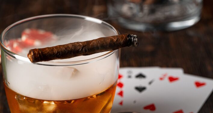 Charuto cubano, copo de uísque e cartas na mesa. Foto que ilustra post sobre Imposto Seletivo (IS). Reprodução: FabrikaPhoto/Envato.