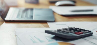 Calculadora com papelada e laptop na mesa no escritório. Foto que ilustra post sobre reforma tributária e Simples Nacional. Reprodução: papans/Envato.