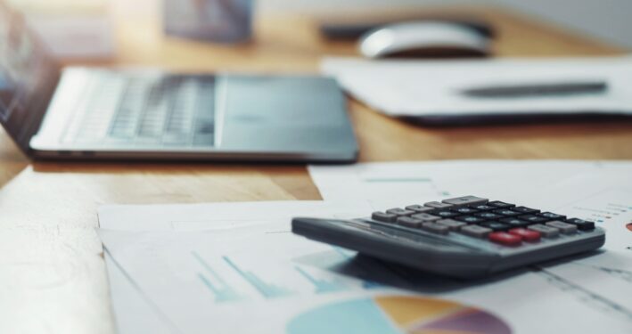 Calculadora com papelada e laptop na mesa no escritório. Foto que ilustra post sobre reforma tributária e Simples Nacional. Reprodução: papans/Envato.
