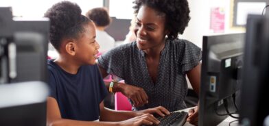 Criança estudando em computador enquanto é auxiliada por professora. Foto que ilustra post sobre IA nas escolas. Reprodução: Envato.
