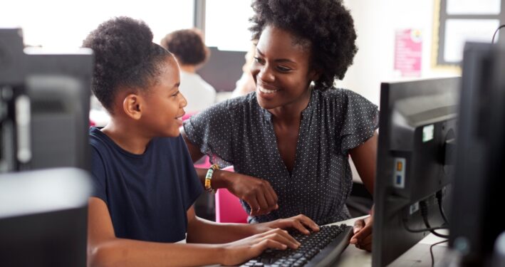Criança estudando em computador enquanto é auxiliada por professora. Foto que ilustra post sobre IA nas escolas. Reprodução: Envato.