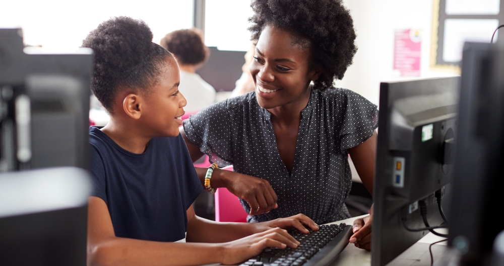 Criança estudando em computador enquanto é auxiliada por professora. Foto que ilustra post sobre IA nas escolas. Reprodução: Envato.